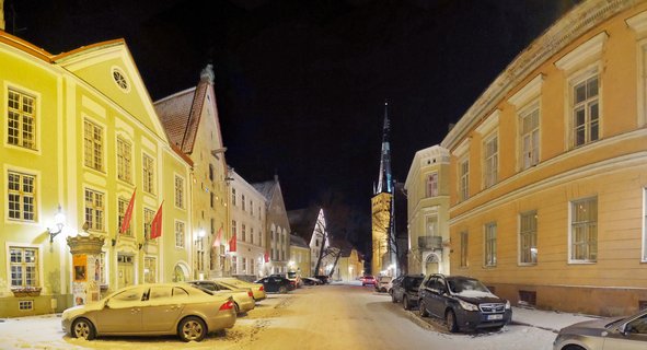 Snow-covered street in Tallinn Old Town at night with illuminated yellow and orange historic buildings, parked cars with snow, and a tall church spire in the distance