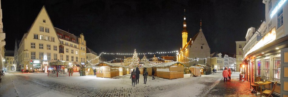 Night view of a Christmas market in a snow-covered medieval square with illuminated wooden stalls, a lit Christmas tree, and historic buildings including a tall tower
