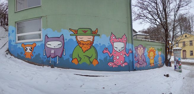 Colorful cartoon character mural painted on green building wall with snow on ground, bare trees, and yellow building in background