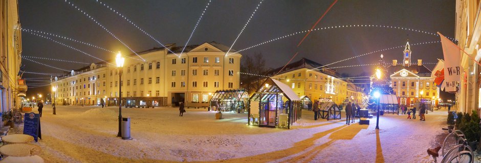 Panoramic night view of Tartu Town Hall Square covered in snow with strings of white lights overhead, illuminated historic buildings, glass market stalls, and people walking through the square