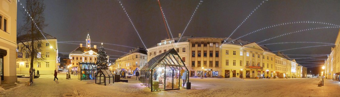 Night view of a snow-covered town square with historic buildings, Christmas tree, and arcing string lights overhead in Tartu, Estonia