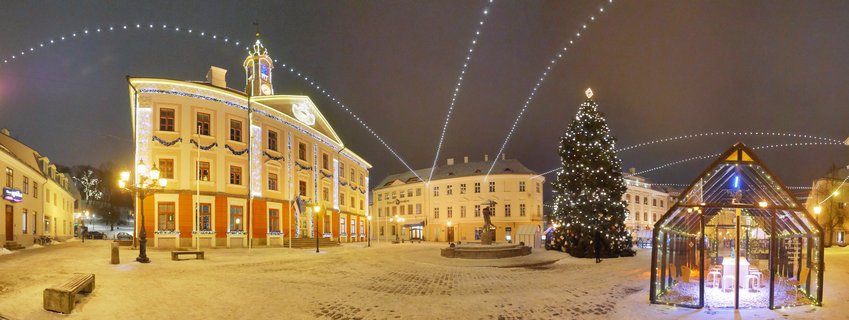 Night view of Tartu Town Hall Square in Estonia during winter, showing the illuminated baroque Town Hall building, decorated Christmas tree with lights, snow-covered cobblestone ground, glass pavilion structure, and festive string lights arching overhead against dark blue evening sky.
