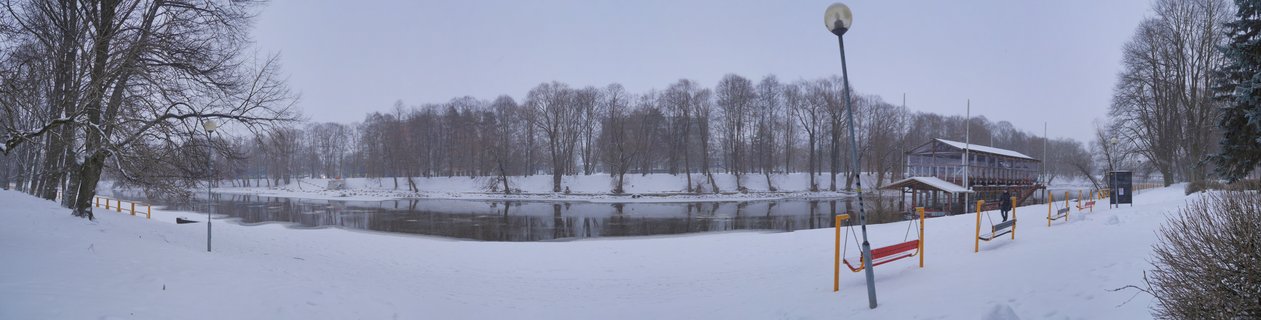 Panoramic winter landscape showing a snow-covered riverbank with bare trees, a riverside building, red benches, and street lamps along the Emajõgi River in Tartu, Estonia