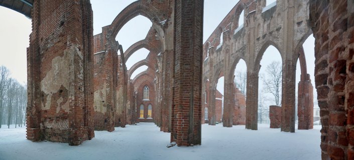 Ruins of a Gothic cathedral with red brick pillars and pointed arches standing in snow-covered ground during winter, bare trees visible through the open structure