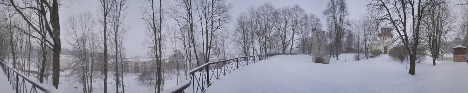 Panoramic winter scene showing snow-covered ground, bare trees, metal railing along elevated walkway, concrete monument sculpture, and water tower building in Tartu, Estonia under gray overcast sky