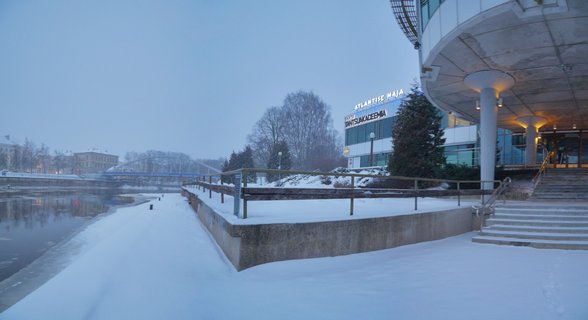 Wide winter landscape showing a partially frozen river in Tartu, Estonia with snow-covered banks, bare trees, buildings in the background, and reflections in dark blue water during twilight