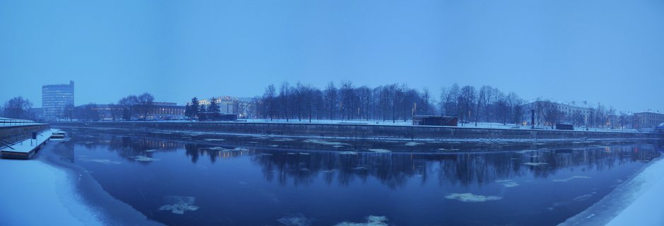 Wide winter landscape showing a partially frozen river in Tartu, Estonia with snow-covered banks, bare trees, buildings in the background, and reflections in dark blue water during twilight