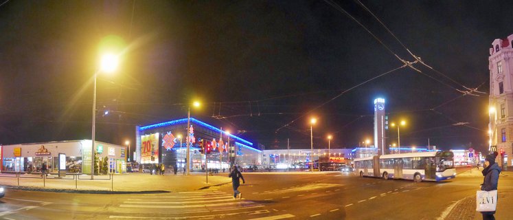 Night panoramic street scene in Riga showing illuminated shopping center with blue Christmas lights, articulated bus, street lights, overhead wires, and pedestrians crossing at intersection with snow on ground