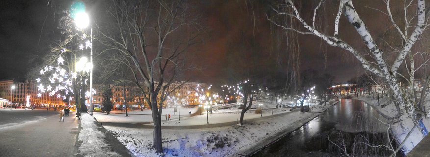 Night panorama of a snow-covered urban park with canal in Riga, Latvia, featuring illuminated star decorations on trees, street lamps, and buildings in the background with people walking on pathways