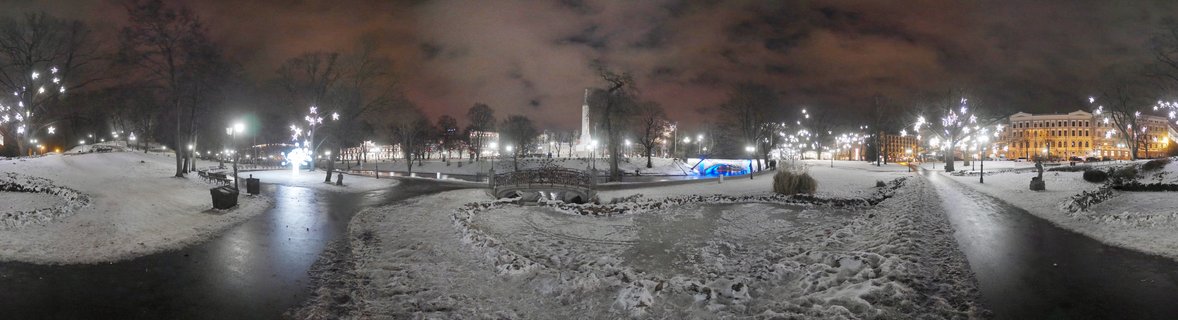 Panoramic night photograph of a snow-covered urban park in Riga, Latvia, with illuminated monument, decorative star lights on trees, and reflective wet pathways
