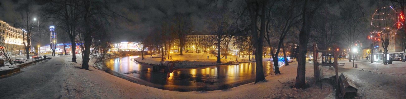 Night panorama of a snow-covered canal in Riga with illuminated buildings, bare trees, street lights reflecting on water, and holiday decorations