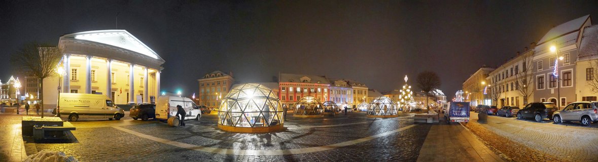 Night view of Vilnius Cathedral Square showing illuminated white geodesic dome structures, historic buildings with classical columns, Christmas tree with lights, cobblestone plaza, parked vehicles, Lithuania