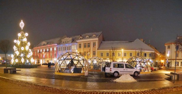 Night photograph of a European town square with a large lit Christmas tree on the left, three glass dome structures in the center, colorful historic buildings in the background, and a white van parked nearby on cobblestone pavement with snow