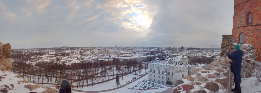 Wide panoramic photograph showing snow-covered Vilnius Old Town cityscape viewed from Gediminas Tower, with historic church spires, baroque buildings, bare trees, and stone fortification walls in foreground under cloudy winter sky