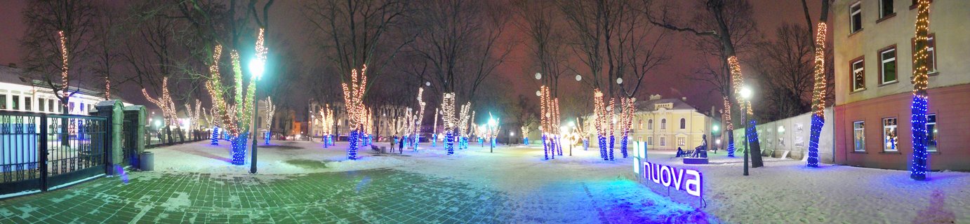Night panoramic view of snow-covered public square with trees wrapped in Christmas lights, blue and white LED decorations, buildings in background, people walking, Kaunas Lithuania