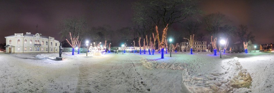 Panoramic night photograph of a snow-covered park with trees decorated in amber and blue Christmas lights, illuminated historical buildings, and street lamps in Kaunas, Lithuania