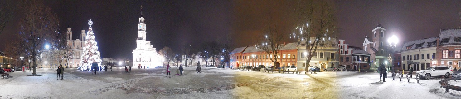 Night view of snow-covered town square with illuminated Christmas tree, historic buildings, church tower, and people walking in Kaunas, Lithuania