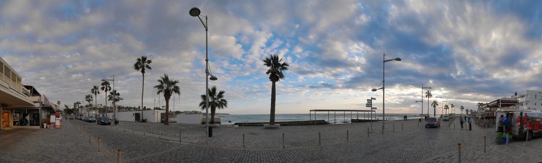 Wide coastal promenade in Paphos, Cyprus with palm trees, cobblestone walkway, street lamps, and Mediterranean Sea under cloudy sky