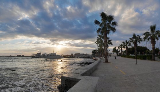 Waterfront promenade in Paphos, Cyprus with palm trees along a paved walkway next to the Mediterranean Sea. Yachts are moored in the harbor under a cloudy sky with the sun setting on the horizon.