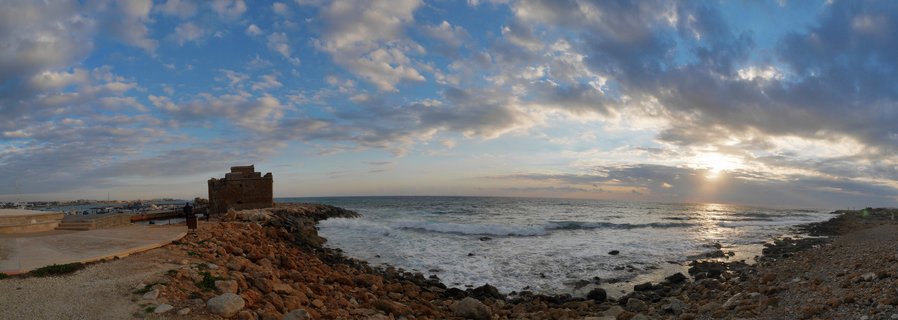 Stone medieval castle on rocky breakwater at Paphos Harbor with sunset over Mediterranean Sea, waves crashing on shore, dramatic cloudy sky with sun rays