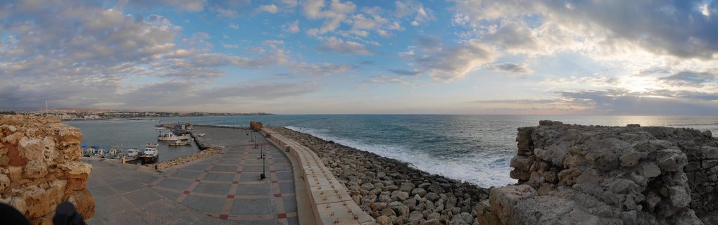 Panoramic coastal view of Paphos harbor in Cyprus showing ancient stone ruins, a paved promenade, docked boats in the marina, breakwater rocks, and the Mediterranean Sea under cloudy sky at sunset