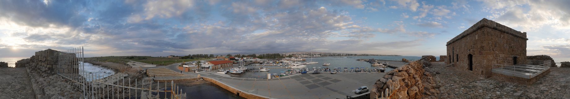 Panoramic view of Paphos Medieval Castle stone fortress overlooking a marina harbor with boats, Mediterranean Sea, and cloudy sky at sunset in Cyprus