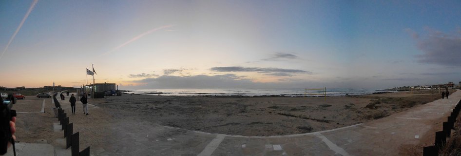 Panoramic beach scene showing sandy shore with volleyball net, paved walkway with pedestrians, flags on poles, parked cars, and Mediterranean Sea at sunset under partly cloudy sky