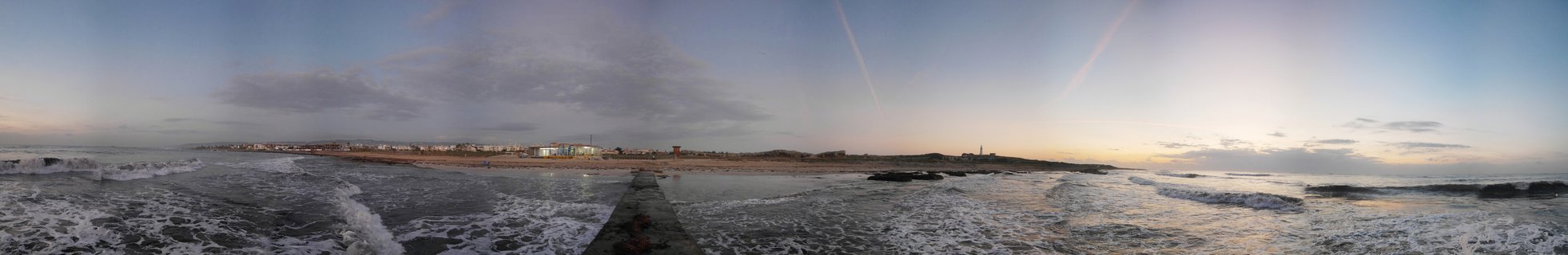 Panoramic view of waves breaking on a beach in Paphos, Cyprus at sunset with a concrete pier extending into the Mediterranean Sea and city buildings visible along the coastline
