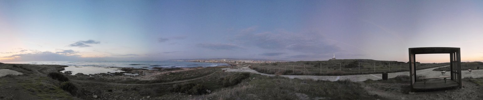Panoramic coastal landscape showing Mediterranean Sea at dusk with rocky shoreline, grassy terrain, wooden shelter structure on the right, and distant city lights visible across the water near Paphos, Cyprus