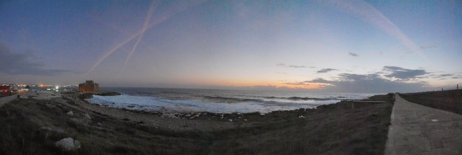 Panoramic view of Paphos Castle on the Mediterranean coast at twilight with waves, rocky shoreline, paved pathway, and sunset colors on the horizon