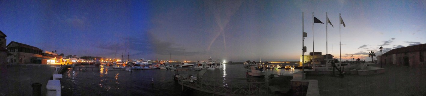 Panoramic view of Paphos harbor at dusk with numerous boats docked in calm water, illuminated buildings along the waterfront, and a deep blue twilight sky