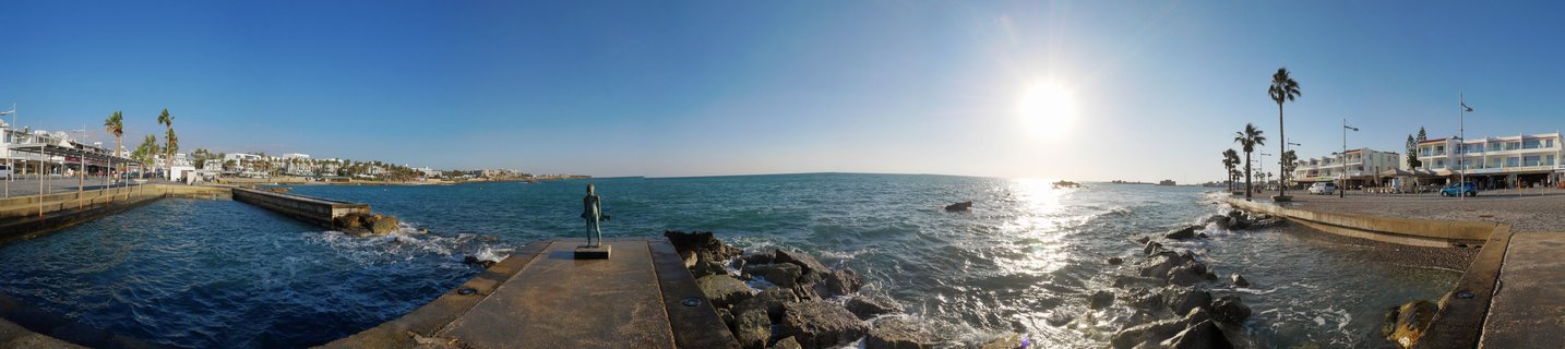 Panoramic coastal scene showing a statue on a pier in Paphos, Cyprus, with turquoise Mediterranean Sea, palm trees, white buildings, and bright sun in clear blue sky