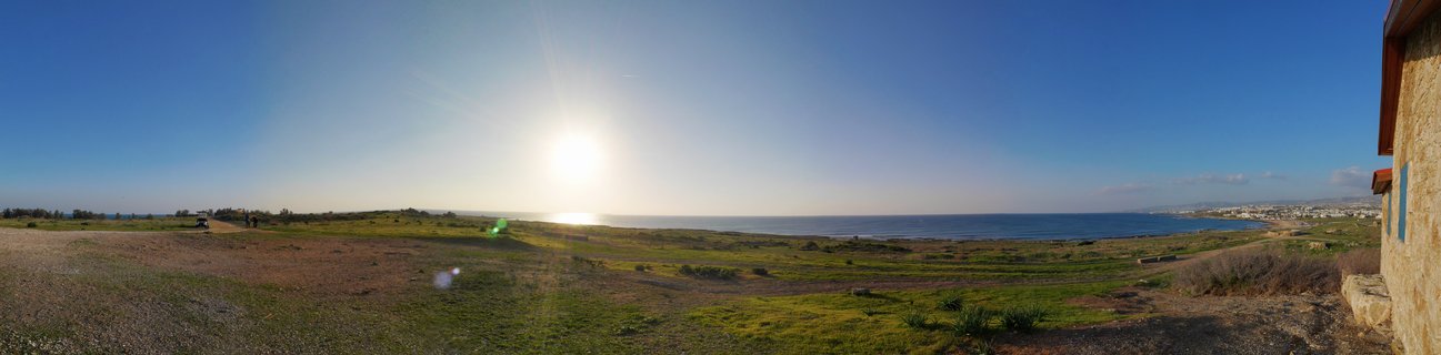 Wide coastal landscape showing grassy terrain, Mediterranean Sea, bright sun in blue sky, and stone building with blue shutters on the right