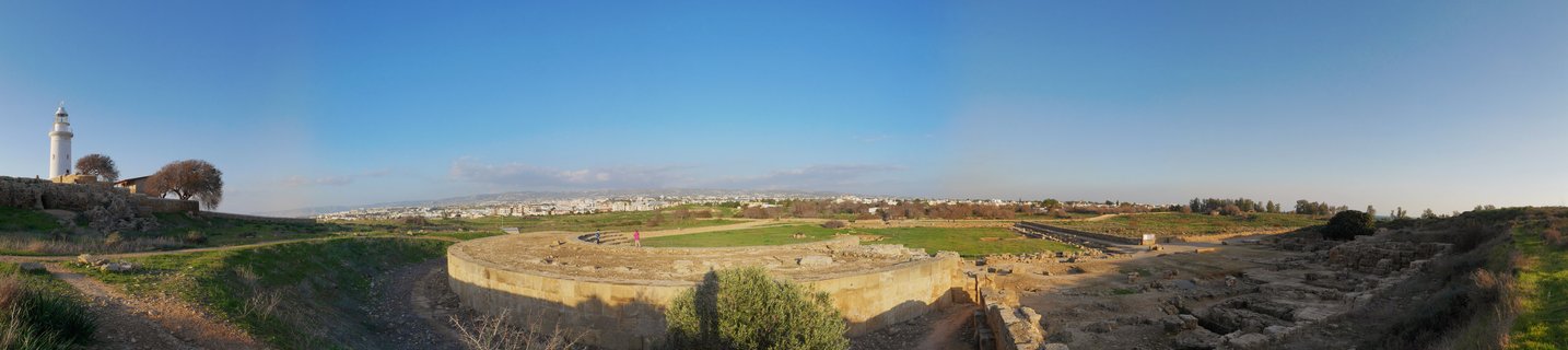 White lighthouse on hillside overlooking ancient stone ruins and archaeological excavation site with city skyline in background under blue sky