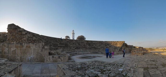 Ancient stone amphitheater ruins with tiered seating, weathered limestone blocks, visitors walking on the orchestra floor, white lighthouse visible in the distance against clear blue sky