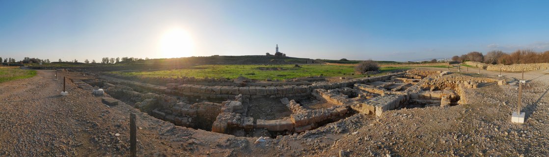 Panoramic view of ancient stone archaeological ruins with gravel pathways and a lighthouse visible on the horizon under a clear blue sky at sunset