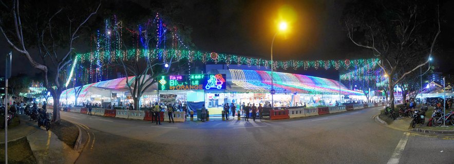 Panoramic night photograph of an outdoor food market in Singapore with colorful string lights hanging above illuminated stalls, people walking on the street, and motorcycles parked along the sides