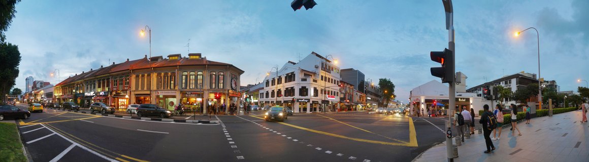 Panoramic street view of Singapore at evening showing traditional shophouses, traffic lights, cars on the road, and pedestrians walking on sidewalks under blue twilight sky