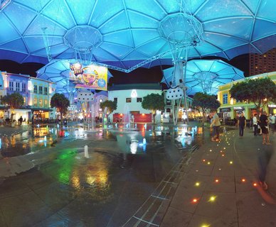 Night view of Clarke Quay plaza in Singapore with blue illuminated canopy structures overhead, water fountains on wet pavement, colorful shophouse buildings, and people walking through the entertainment district