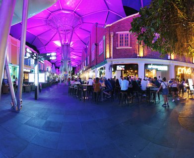 Night scene at Clarke Quay Singapore showing outdoor dining area beneath purple and magenta LED-lit geometric canopy structures. People seated at high tables and stools along riverside promenade. Historic red brick warehouse buildings with restaurant signage visible in background. Steel support columns and artificial lighting throughout.
