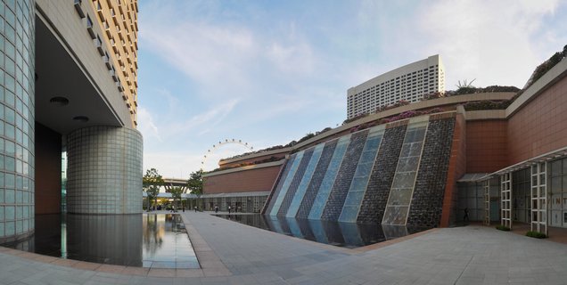 Modern plaza in Singapore featuring a cascading water wall with glass panels, reflective pools, tall buildings, and the Singapore Flyer wheel visible in the background under a blue sky with scattered clouds
