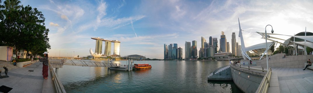 Panoramic view of Singapore's Marina Bay showing Marina Bay Sands hotel, ArtScience Museum, and CBD skyscrapers across the water with a tourist boat and waterfront promenade in the foreground