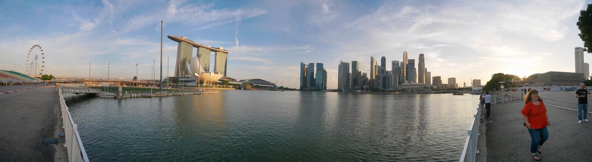 Panoramic view of Singapore Marina Bay showing Marina Bay Sands hotel, ArtScience Museum, Singapore Flyer wheel, and downtown skyscrapers across calm water with pedestrians on waterfront promenade