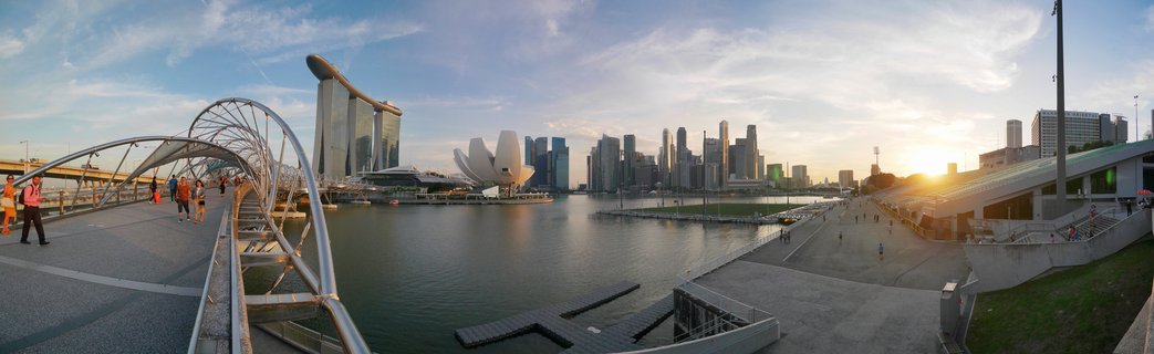 Panoramic cityscape of Singapore Marina Bay at sunset showing the Helix Bridge with pedestrians, Marina Bay Sands hotel, ArtScience Museum, and downtown skyline across the water