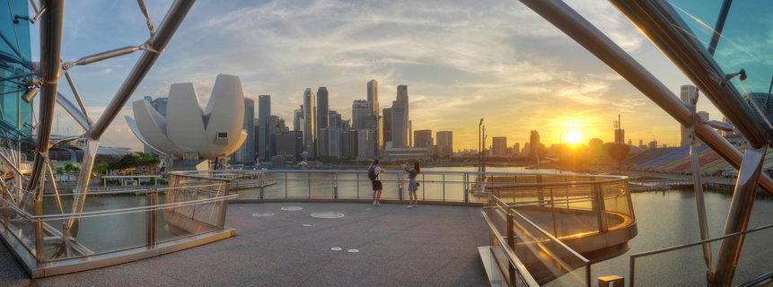 Panoramic view of Singapore's Marina Bay skyline at sunset from Helix Bridge, showing ArtScience Museum, skyscrapers, and two tourists on the viewing platform