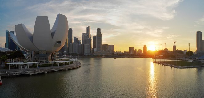 Panoramic view of Singapore Marina Bay at sunset showing the white lotus-shaped ArtScience Museum on the left, skyscrapers of the CBD skyline in the center background, and golden sunlight reflecting on the bay waters