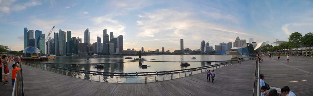 Panoramic view of Singapore's Marina Bay showing the CBD skyline with skyscrapers, ArtScience Museum, waterfront promenade with people walking on wooden boardwalk, and bay water with floating barriers under partly cloudy sky