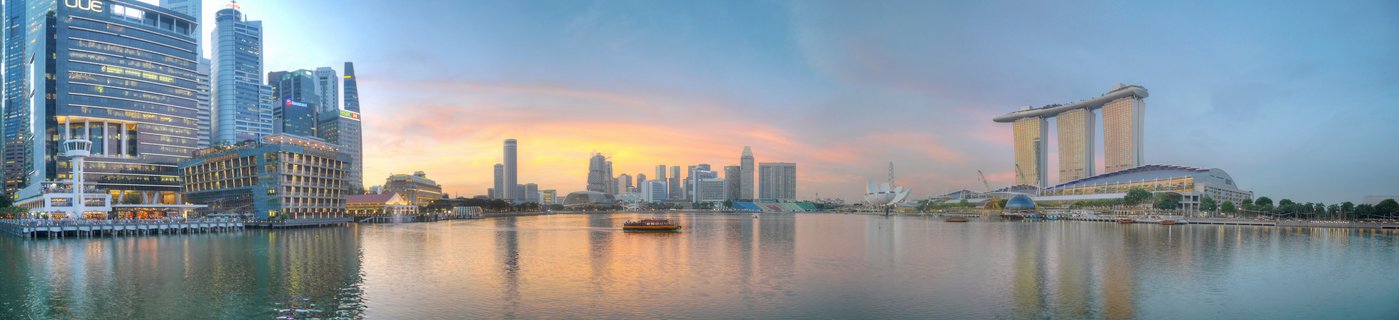 Panoramic view of Singapore Marina Bay skyline at sunset with Marina Bay Sands hotel, CBD skyscrapers, waterfront promenade, and boat on calm water with sky reflections