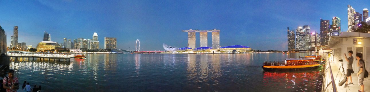 Panoramic view of Singapore Marina Bay at dusk showing illuminated skyscrapers including Marina Bay Sands, Singapore Flyer wheel, waterfront boats, and city lights reflecting on water with people viewing from promenade