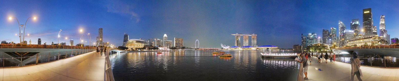 Panoramic view of Singapore's Marina Bay at dusk showing illuminated skyscrapers including Marina Bay Sands, Singapore Flyer wheel, waterfront promenade with pedestrians, and tourist boats on the water with city lights reflecting on the bay surface under a deep blue twilight sky